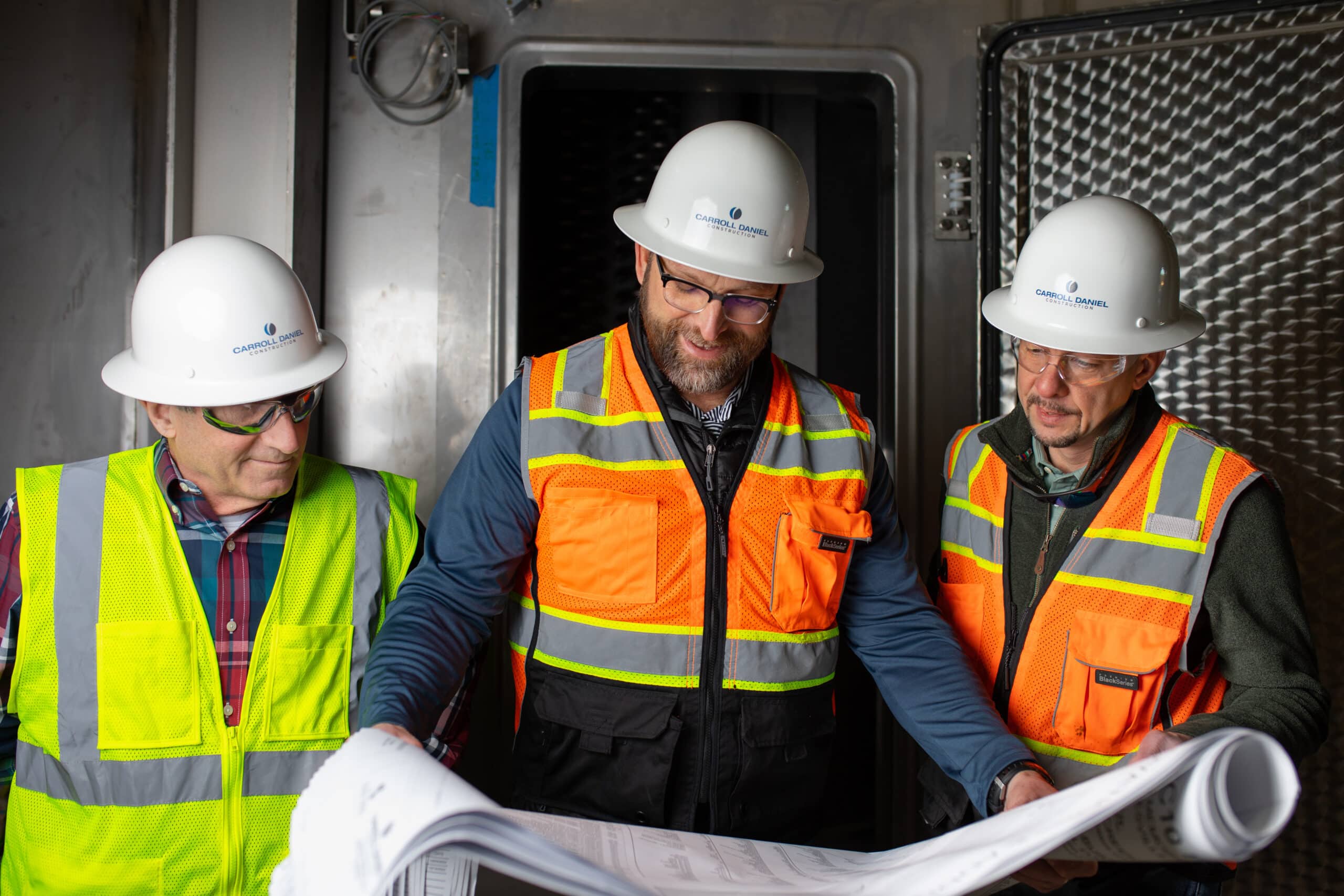 three men in hard hats, safety vest, and safety goggles look over a set of architectural plans